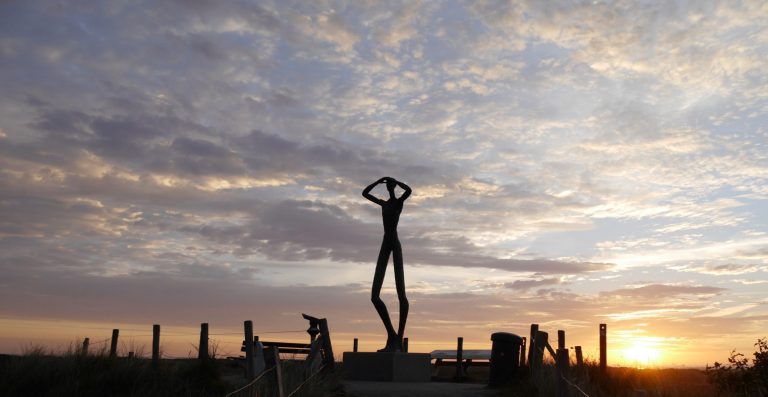 Eine menschliche Statue auf einem Aussichtspunkt hält Arme ans Gesicht und schaut  in die Ferne. Dahinter ist ein Sonnenaufgang zu sehen.